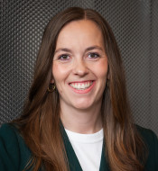 Frau mit langen, braunen Haaren lächelt in dunkelgrünem Blazer vor metallischer, strukturierter Oberfläche.