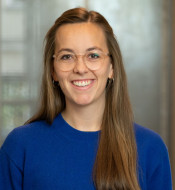 Pauline Bombeck stands in a bright room, wearing a blue sweater and glasses, smiling with an open posture.