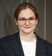 Mitarbeiterfoto: Friederike Micheael, Person mit kurzen, hellen Haaren, gestreiftem Blazer und Brille vor metallischer Wand.