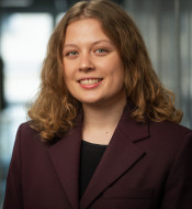 Junge Frau mit lockigem, schulterlangem Haar in dunkelviolettem Blazer, lächelt vor unscharfem, hellem Hintergrund.