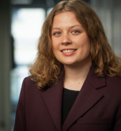 Junge Frau mit lockigem, schulterlangem Haar in dunkelviolettem Blazer, lächelt vor unscharfem, hellem Hintergrund.