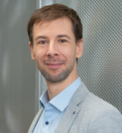 Mann mit braunen Haaren und Bart in hellblauem Hemd und grauem Blazer vor metallischer Wand.