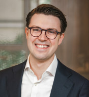 Person mit kurzen, dunklen Haaren und Brille, in hellem Hemd und dunklem Blazer, lächelt vor unscharfem Hintergrund.