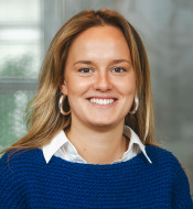 Person mit langen, hellen Haaren, blauem Pullover, weißem Hemd und runden Ohrringen, lächelt vor unscharfem Hintergrund.