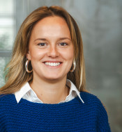 Person mit langen, hellen Haaren, blauem Pullover, weißem Hemd und runden Ohrringen, lächelt vor unscharfem Hintergrund.