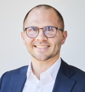 Person mit Brille lächelt in die Kamera, trägt dunklen Anzug und weißes Hemd, neutraler Hintergrund.