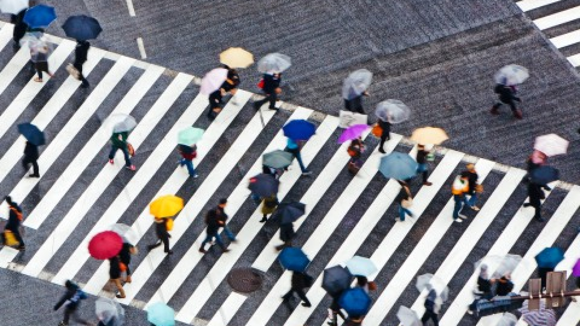 Das Bild zeigt eine belebte Straßenkreuzung mit mehreren Zebrastreifen, auf denen zahlreiche Menschen mit Regenschirmen unterwegs sind. 