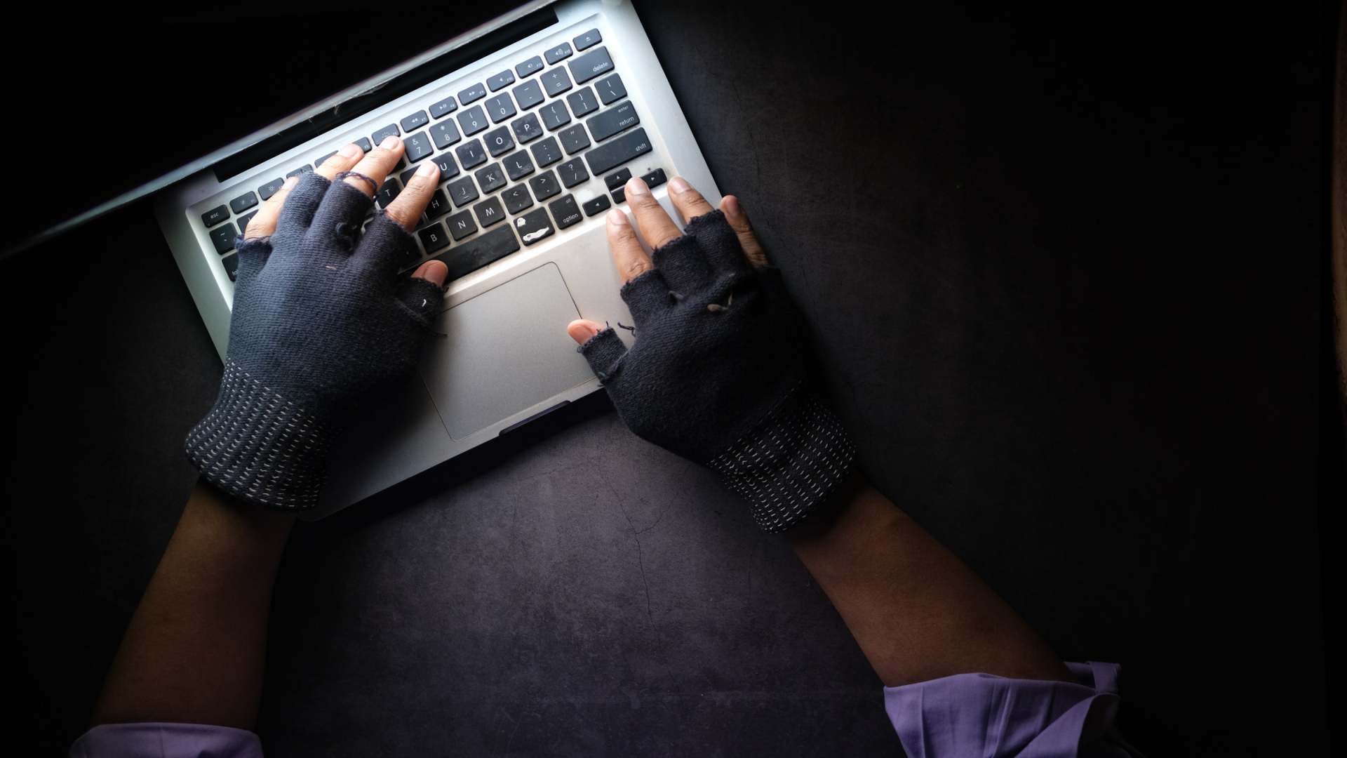  Hände in fingerlosen Handschuhen tippen am Laptop, dunkle Atmosphäre 