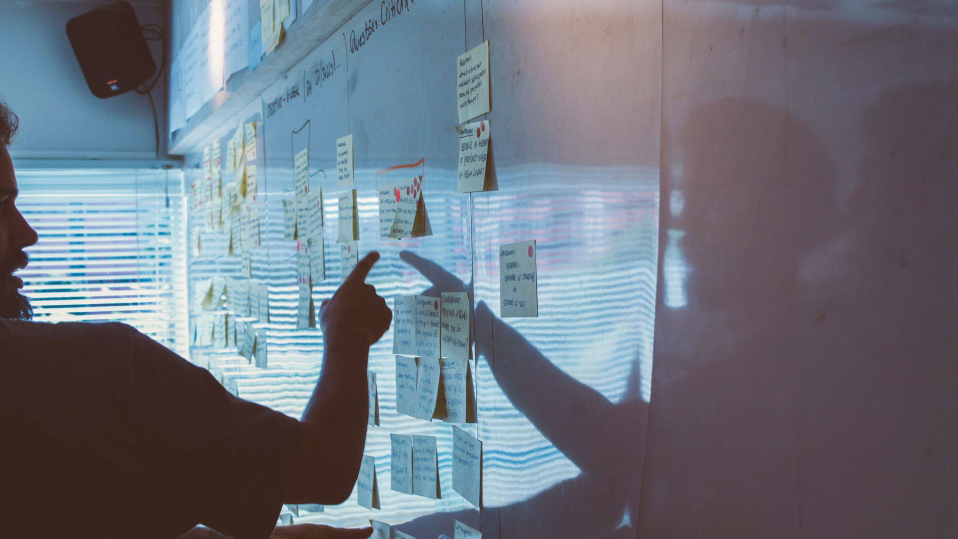 Person zeigt auf Haftnotiz an Whiteboard, umgeben von bunten Notizen und Licht durch Fenster.