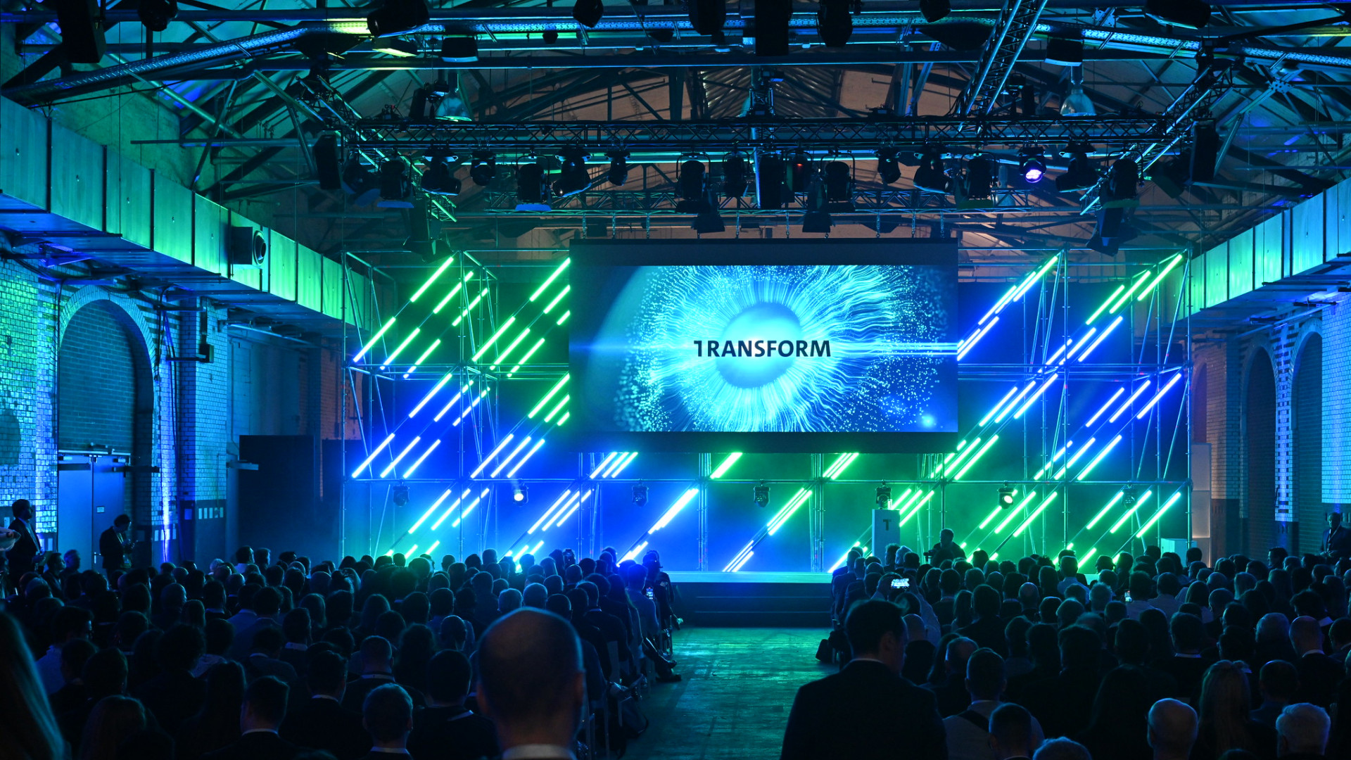 Publikum in dunkler Business-Kleidung sieht in einem Saal zu einer Bühne auf, wo ein Banner mit dem Wort "TRANSFORM" und einem sternähnlichen visuellen Effekt präsentiert wird. Die Szene ist in blaues und grünes Licht getaucht.