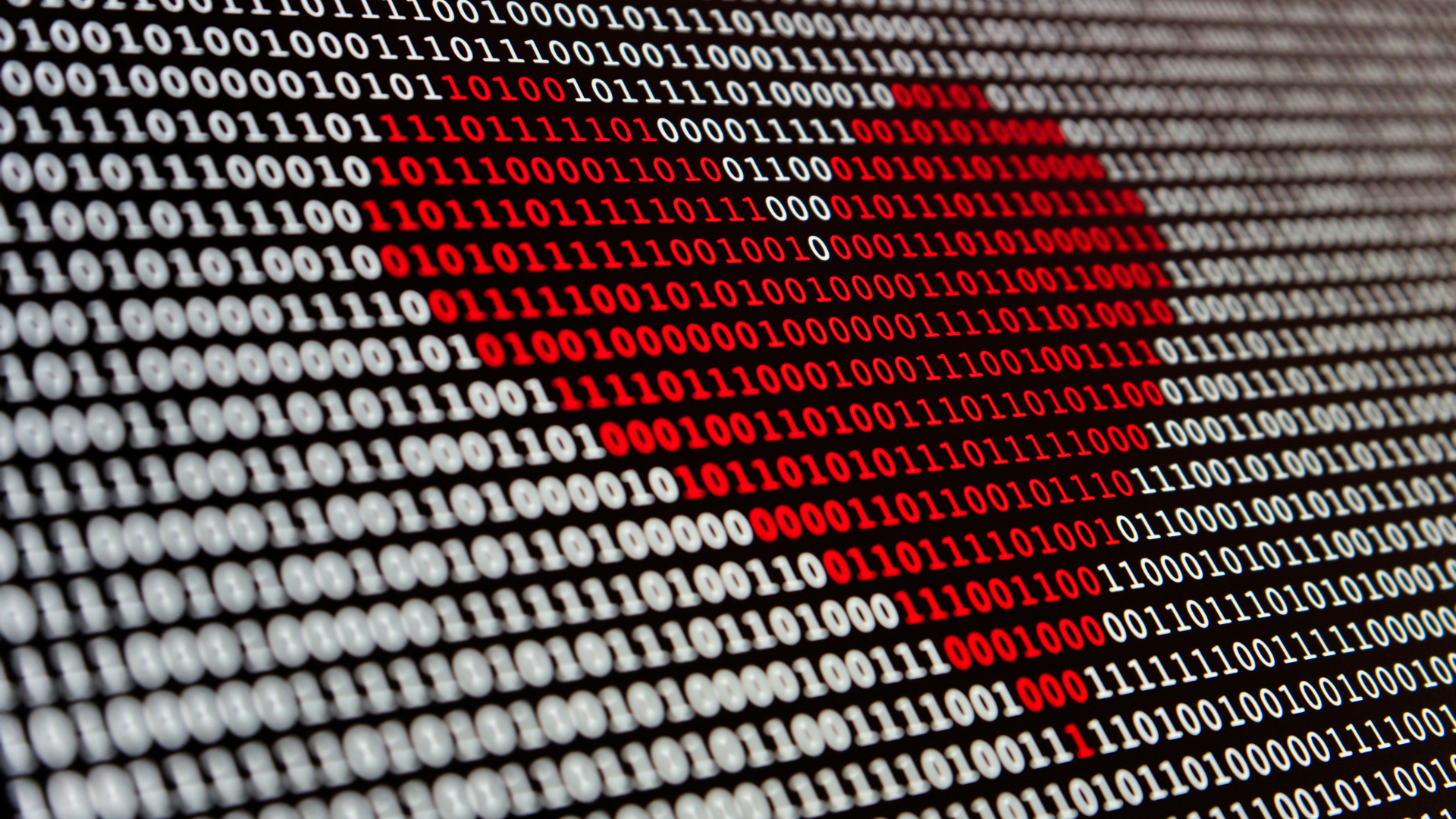 Das Bild zeigt eine großformatige Darstellung von binärem Code, bestehend aus den Zahlen 0 und 1. In der Mitte formt der Code ein rotes Herz, das sich von den umliegenden schwarzen und weißen Einsen und Nullen abhebt. Die Zahlen sind in Reihen angeordnet, wobei das Herz als optisches Motiv heraussticht. Der Hintergrund ist dunkel, während der Code hell und klar lesbar ist.