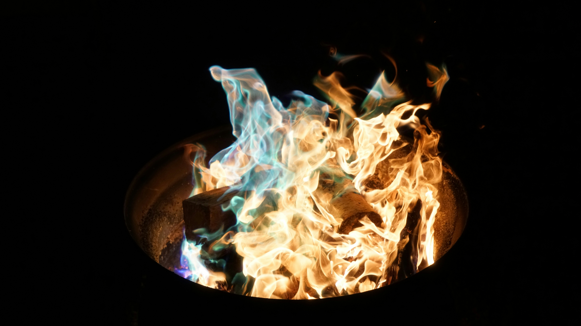 In der Mitte des Bildes lodert ein Feuer in einer dunklen Umgebung. Die Flammen sind teils orange, teils leuchtend blau-grün, was auf eine besondere chemische Reaktion oder Brennstoffe hinweist. Holzscheite liegen im Feuer und werden von den Flammen umschlossen. Das Feuer befindet sich in einem runden Metallbehälter, der nur teilweise im Bild zu sehen ist. Die Umgebung ist ansonsten dunkel und es gibt keine weiteren sichtbaren Objekte.