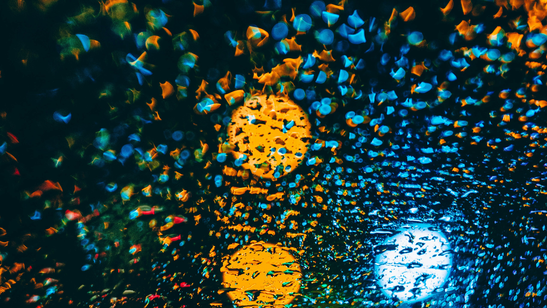 Eine Nahaufnahme von Regentropfen auf einer Fensterscheibe bei Nacht, die bunte Lichter von Straßenlaternen und Fahrzeugen im Hintergrund reflektieren. Die Lichter erscheinen verschwommen und in leuchtenden Farben wie Gelb, Blau, Rot und Grün, die durch die Tropfen in bokehartigen Mustern verzerrt werden. Das Bild vermittelt eine atmosphärische, abstrakte Stimmung mit einem starken Fokus auf Licht- und Farbkontraste.