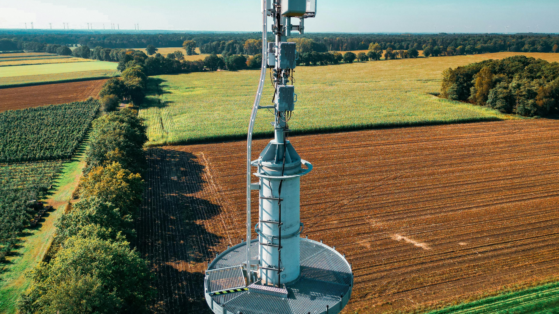 Die Fotografie zeigt einen Mobilfunkmast in einer ländlichen Umgebung. Der Mast steht zentral im Bild und ist mit mehreren Antennen und technischen Geräten ausgestattet. Im Hintergrund erstreckt sich eine weite, landwirtschaftlich genutzte Fläche mit verschiedenen Feldern, die von Baumreihen begrenzt sind. Der Himmel ist klar und türkisblau, was auf sonniges Wetter hinweist. Am Horizont sind Windkraftanlagen erkennbar, die in die Landschaft eingebettet sind.