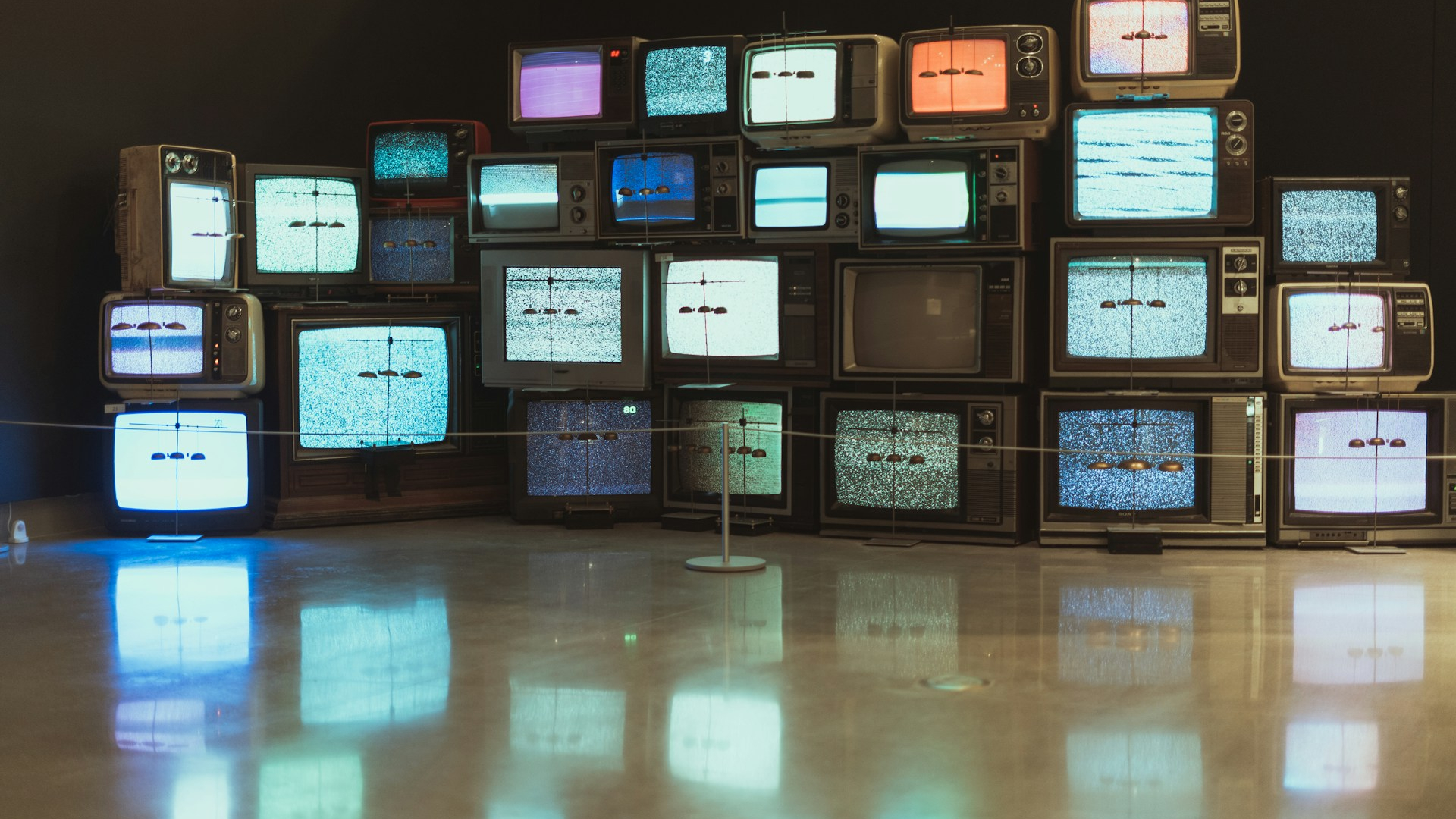Eine Kunstinstallation aus mehreren Dutzend gestapelten Vintage-Röhrenfernsehern in einem dunklen Raum. Die Geräte sind in verschiedenen Größen und Designs gehalten, viele mit Drehknöpfen und Holzrahmen. Die Bildschirme zeigen statisches Rauschen oder farbige, verzerrte Bilder in Blau, Rot und Grün. Einige Fernseher sind ausgeschaltet. Vor der Installation sind feine Drähte mit kleinen Gewichten gespannt. Der glänzende Boden spiegelt die Lichter der Bildschirme.