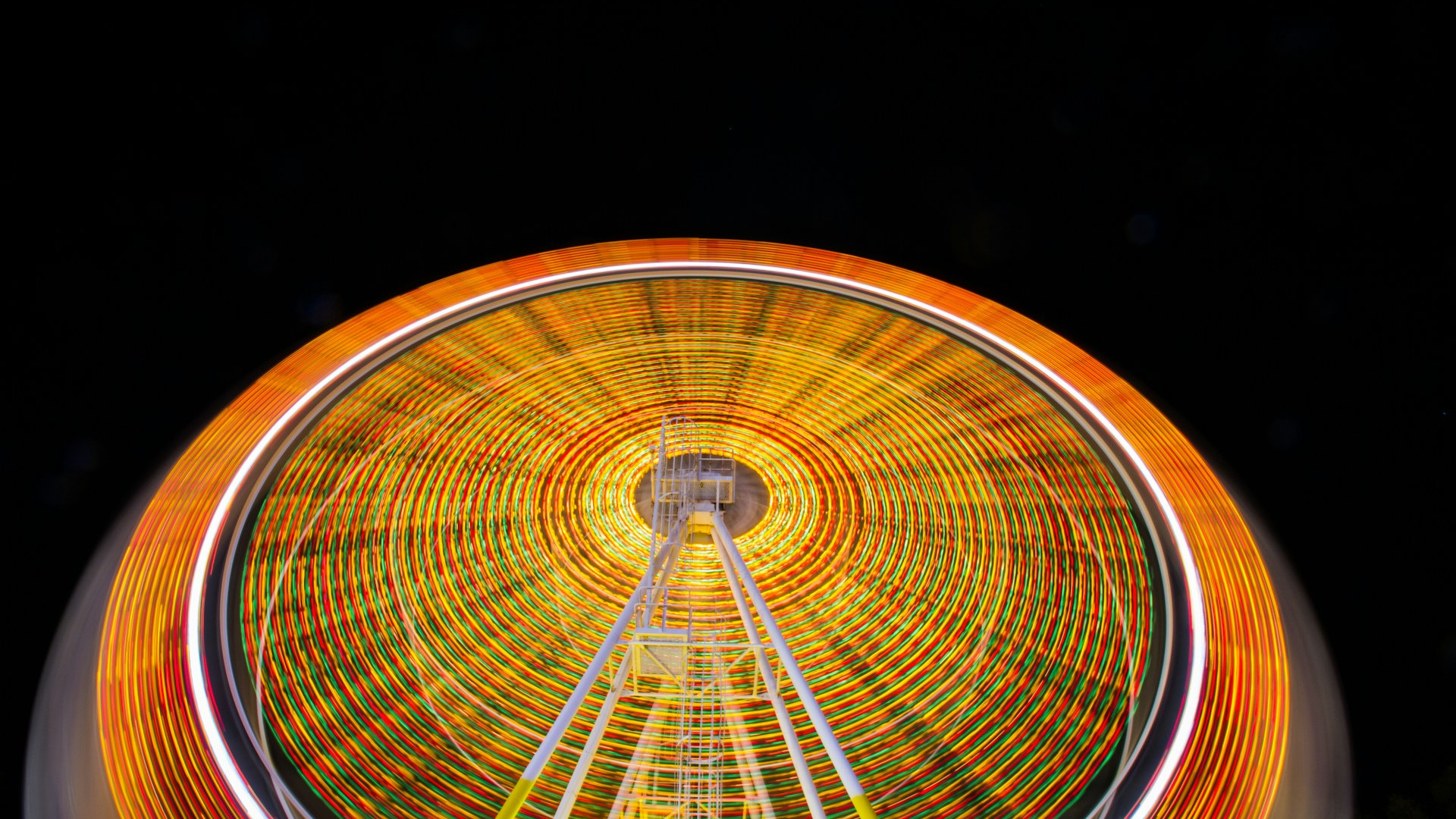 Ein großes Riesenrad dreht sich in der Dunkelheit der Nacht. Durch die Langzeitbelichtung entsteht ein faszinierender Lichteffekt: Die bunten LED-Leuchten des Riesenrads formen konzentrische Kreise in Gelb-, Rot-, Grün- und Weißtönen. Die Konstruktion des Riesenrads mit gelben und weißen Stützen ist in der unteren Bildhälfte zu sehen, während der dunkle Nachthimmel den leuchtenden Effekt verstärkt.