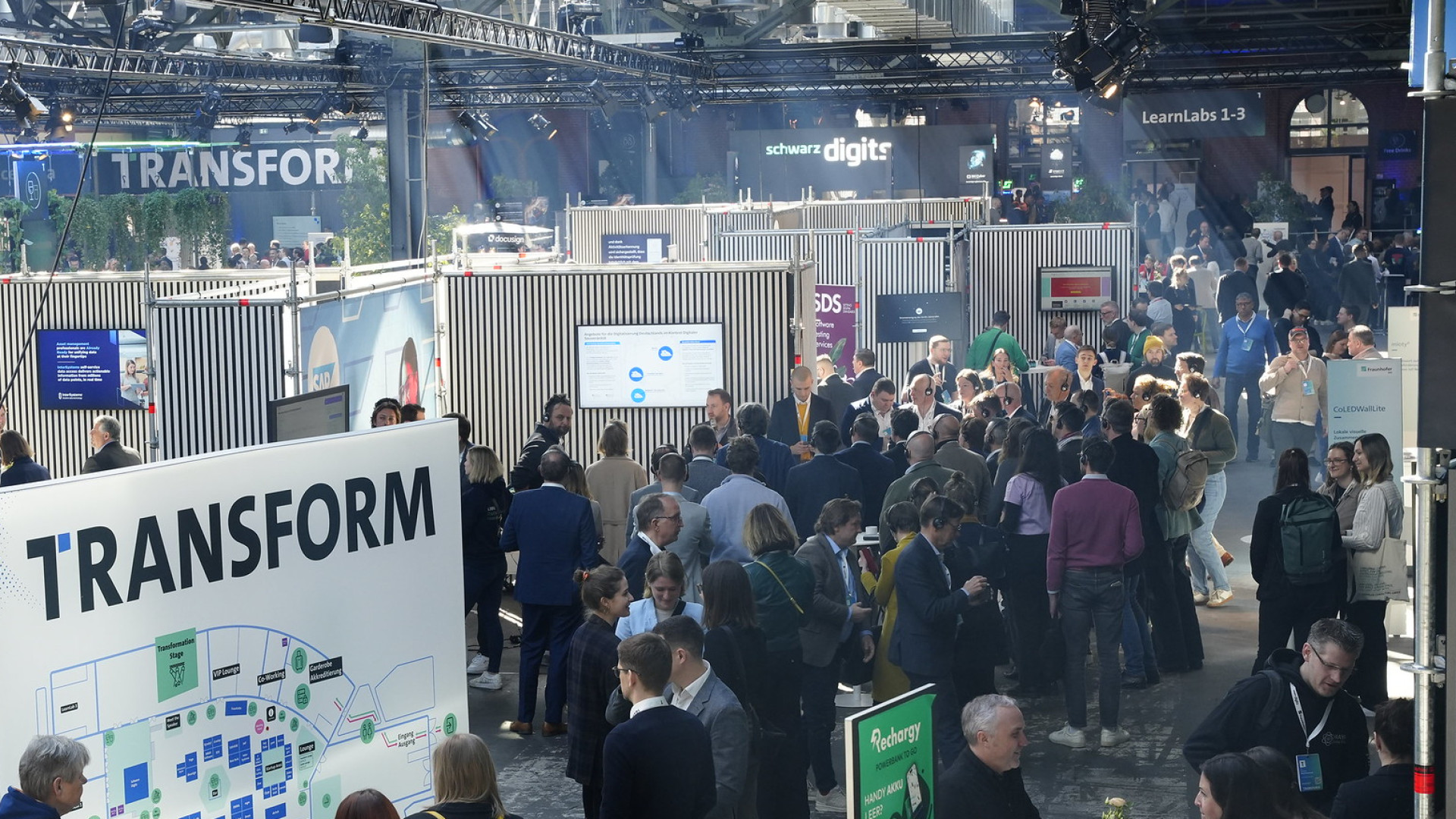 Die STATION Berlin ist gefüllt mit zahlreichen Menschen, die sich in Gruppen unterhalten oder durch die Gänge zwischen den Ständen bewegen. Im Vordergrund steht eine große Infotafel mit der Aufschrift „TRANSFORM“ und einer Übersichtskarte der Veranstaltung. Mehrere Personen betrachten diese Tafel. Im Hintergrund befinden sich zahlreiche Messestände mit Bildschirmen und Bannern, während Besucher geschäftig netzwerken. Die Halle ist industriell gestaltet mit hohen Decken und Stahlträgern.