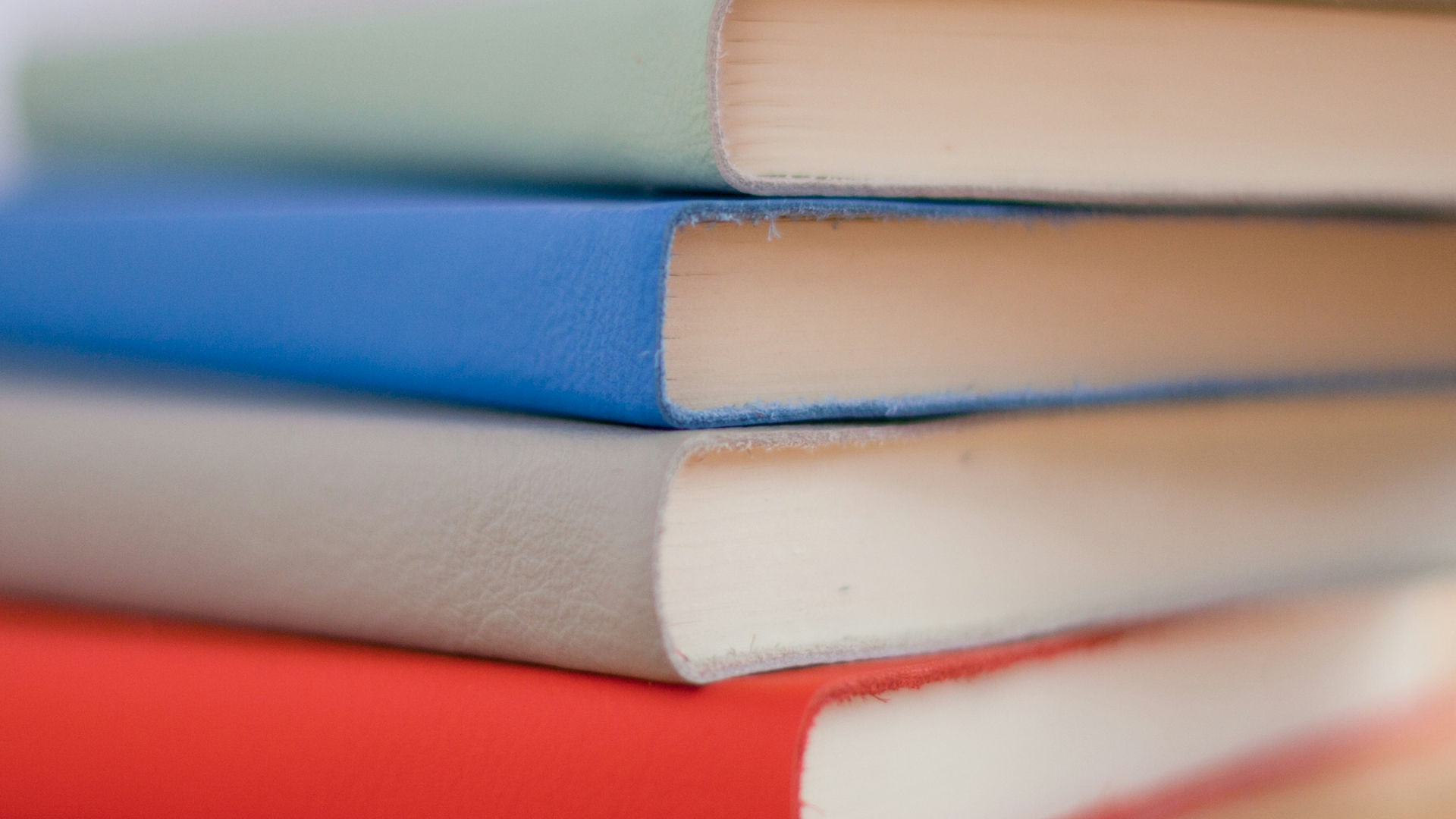 Vier geschlossene Notizbücher liegen horizontal gestapelt auf einem hellen Holztisch. Die Einbände sind glatt und unifarben, von oben nach unten in Hellgrün, Blau, Grau und Rot. Die Bücher sind gleich groß und gleichmäßig übereinandergelegt, mit den Schnittkanten der Seiten nach vorn gerichtet. Der Hintergrund ist unscharf in hellen Rosatönen gehalten, ohne erkennbare Details. Keine Personen im Bild.