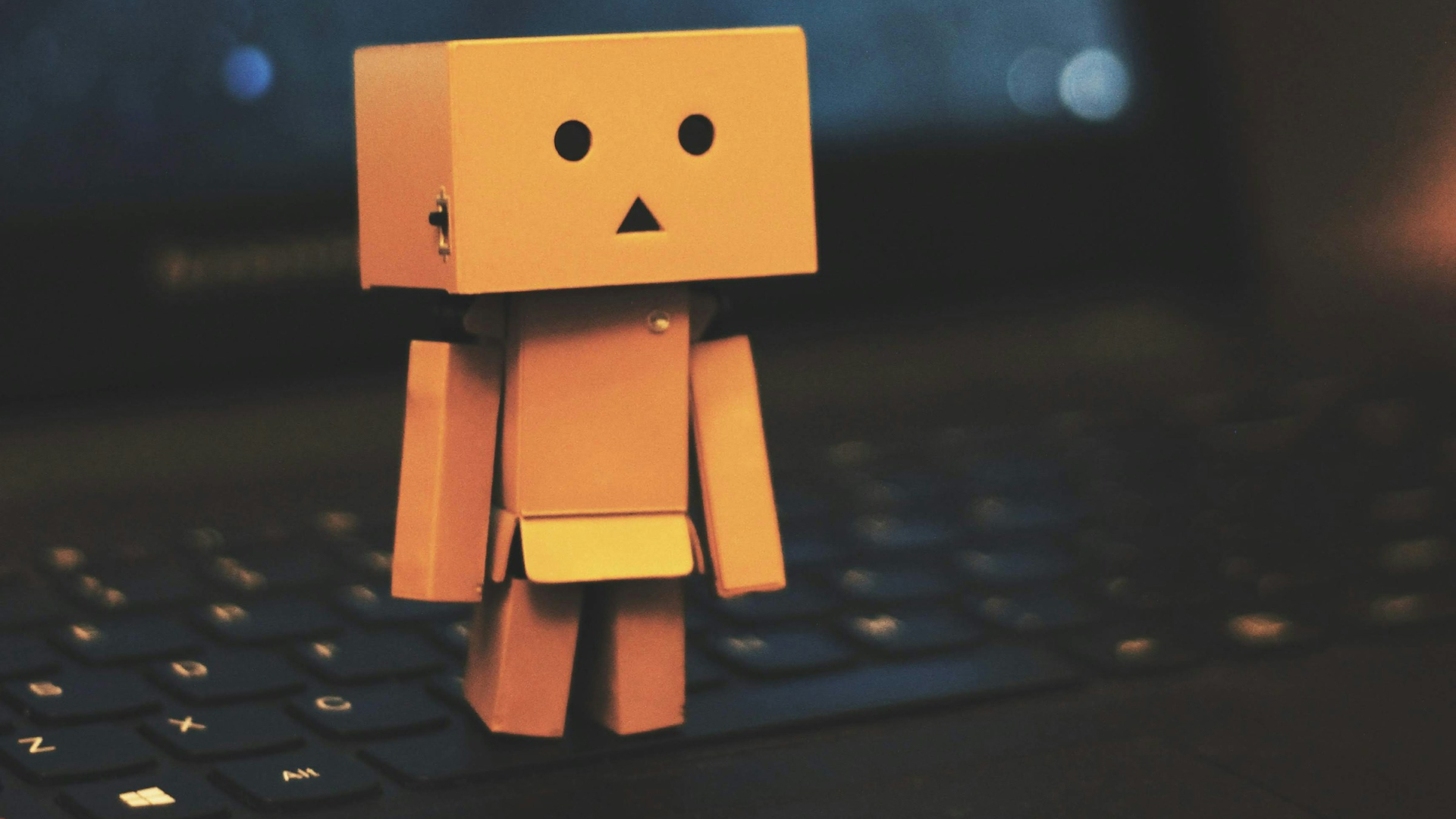 Eine kleine Figur aus Pappkarton mit einem eckigen Kopf, zwei runden Augen und einem dreieckigen Mund steht auf der Tastatur eines Laptops. Ihre Arme und Beine sind beweglich. Der Laptop ist schwarz, und sein Bildschirm im Hintergrund zeigt eine verschwommene, dunkle Ansicht mit blauen und orangen Lichtreflexen. Die Szene ist warm beleuchtet, wodurch die Figur und die Tastatur weiche Schatten werfen.