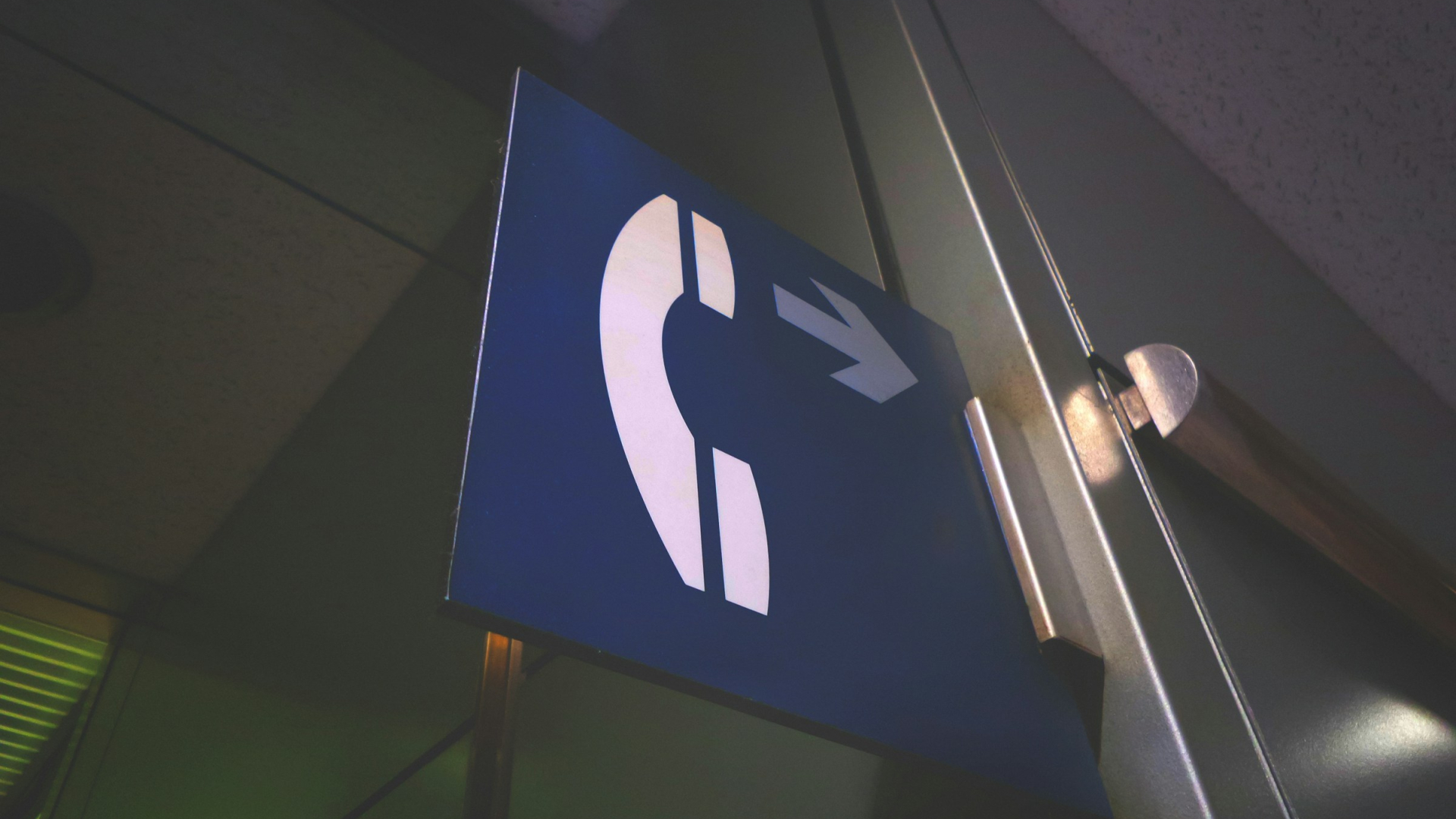 Hinweisschild mit Telefonhörer-Symbol und Pfeil nach rechts, montiert an einer Wand in einem schwach beleuchteten Innenraum.