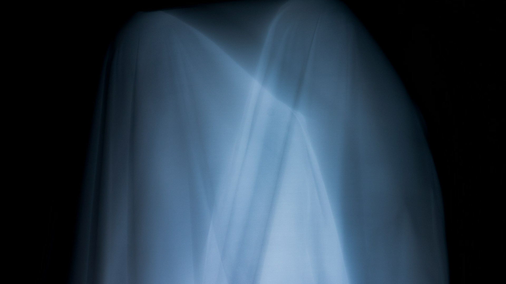 Eine Person steht in einem dunklen Raum und ist vollständig mit einem dünnen, hellen Stoff bedeckt. Der Stoff wirkt durchscheinend und leicht glänzend, wobei weiche Falten und diagonale Linien sichtbar sind. Die Konturen der Person sind unter dem Tuch nur schemenhaft erkennbar, was der Szene ein geheimnisvolles, fast geisterhaftes Erscheinungsbild verleiht. Es sind keine weiteren Gegenstände oder Personen im Bild zu sehen. Es kann symbolisch für Ghosting stehen.