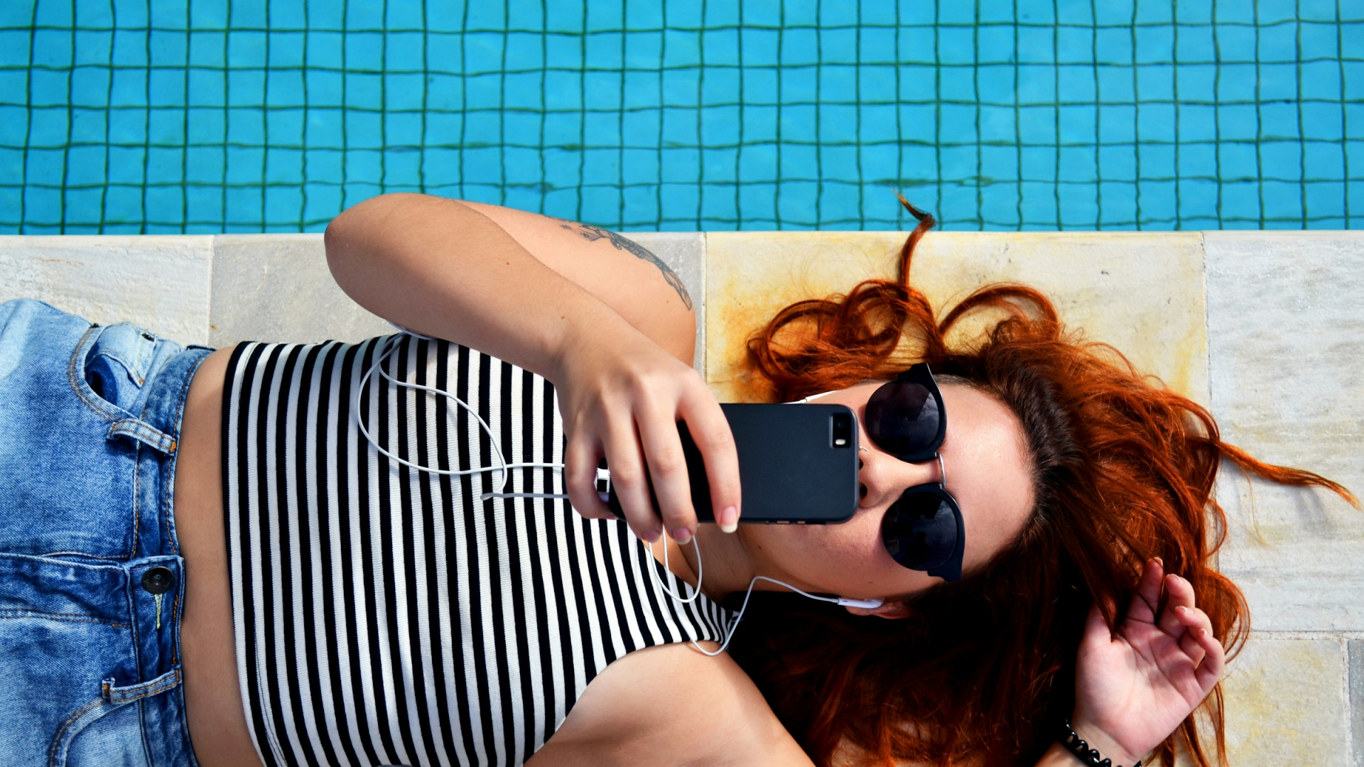 Eine junge Frau liegt am Rand eines Pools und hält ein schwarzes Smartphone direkt vor ihrem Gesicht.