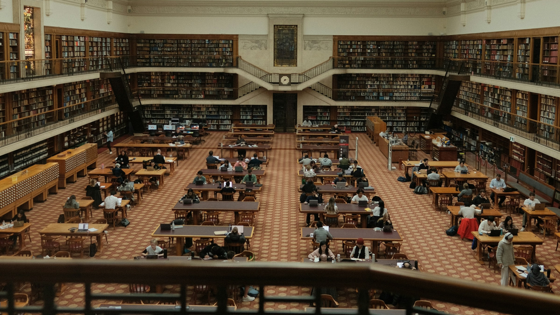 In einer großen, historischen Bibliothek mit hoher Decke sitzen zahlreiche Studierende an langen Holztischen. 