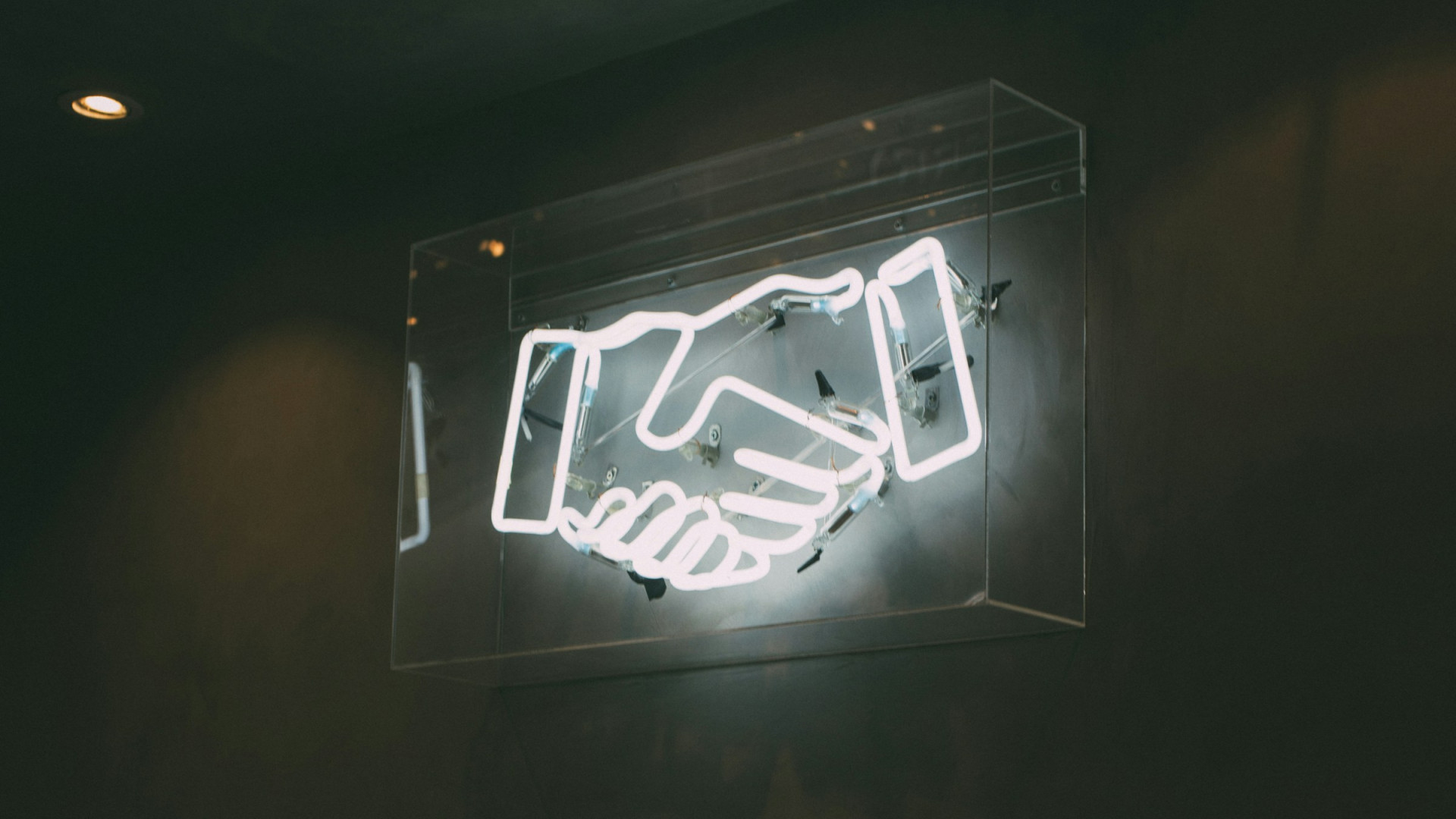 In einem dunklen Raum ist ein leuchtendes Neon-Symbol an einer Wand montiert. Das Symbol zeigt zwei stilisierte Hände im Handschlag.