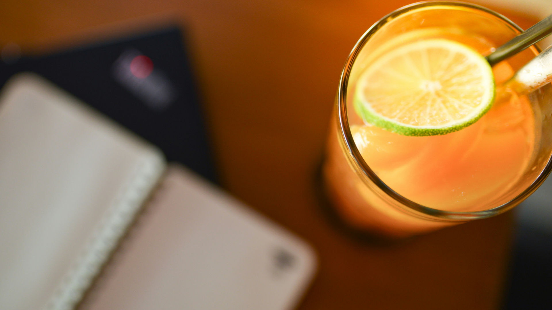 Auf einem Holztisch steht ein Glas mit orangefarbenem Getränk, Eiswürfeln und einer Limettenscheibe.