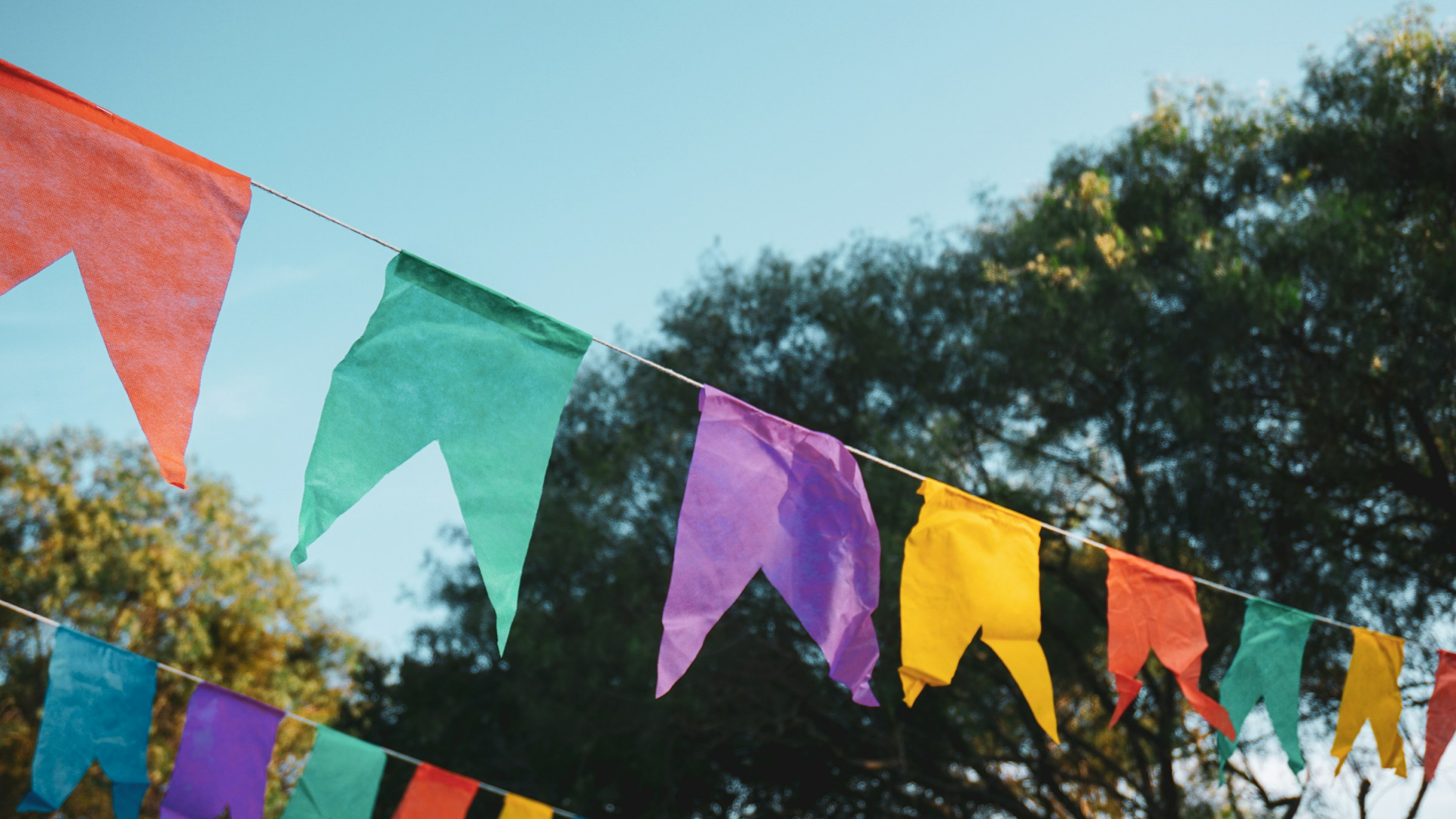 Bunte Wimpelgirlanden hängen im Freien vor blauem Himmel und grünen Bäumen.