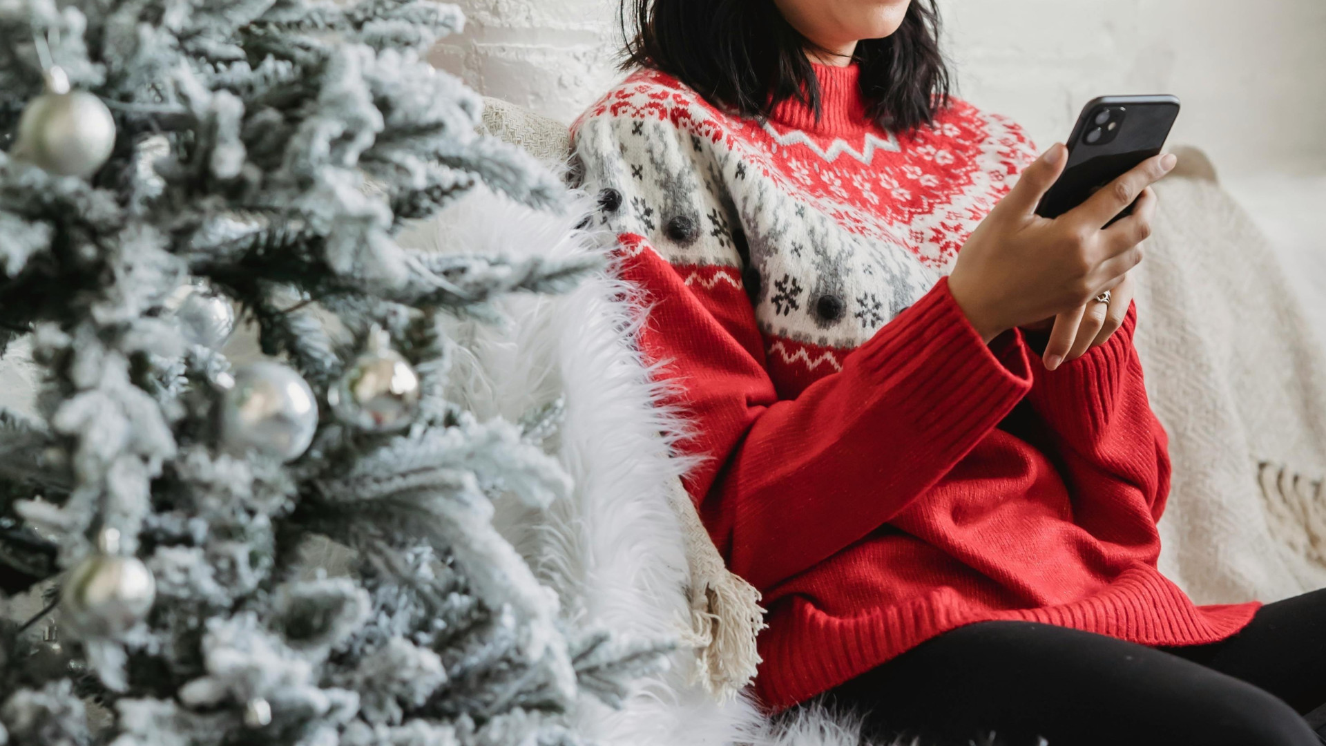 Person mit Weihnachtspullover sitzt neben einem Weihnachtsbaum und Geschenken und hält dabei ein Smartphone in der Hand.