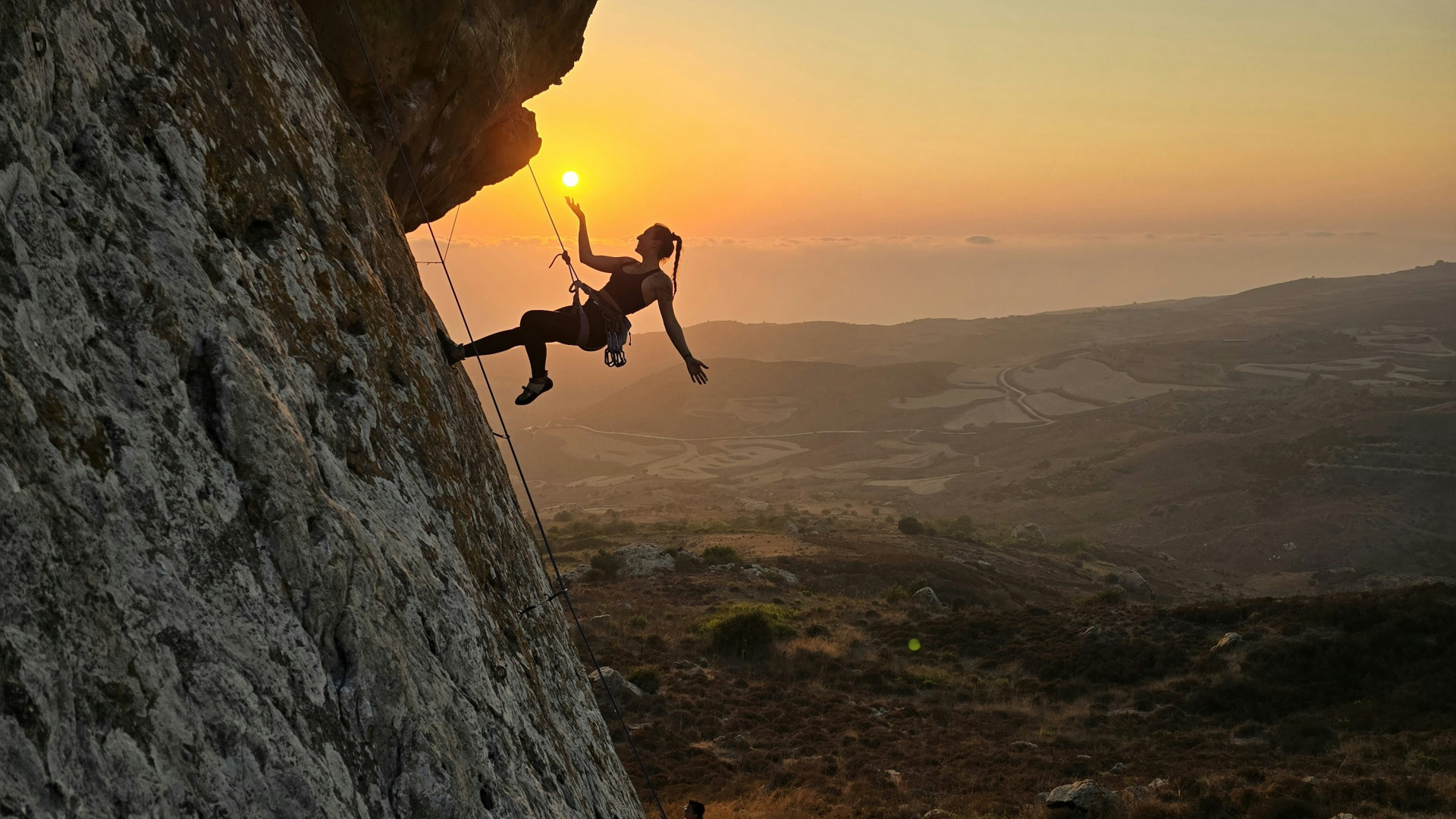 Frau klettert an einer Felswand, zwei Personen beobachten sie. Klettermatten liegen am Boden, Sonnenaufgang im Hintergrund.