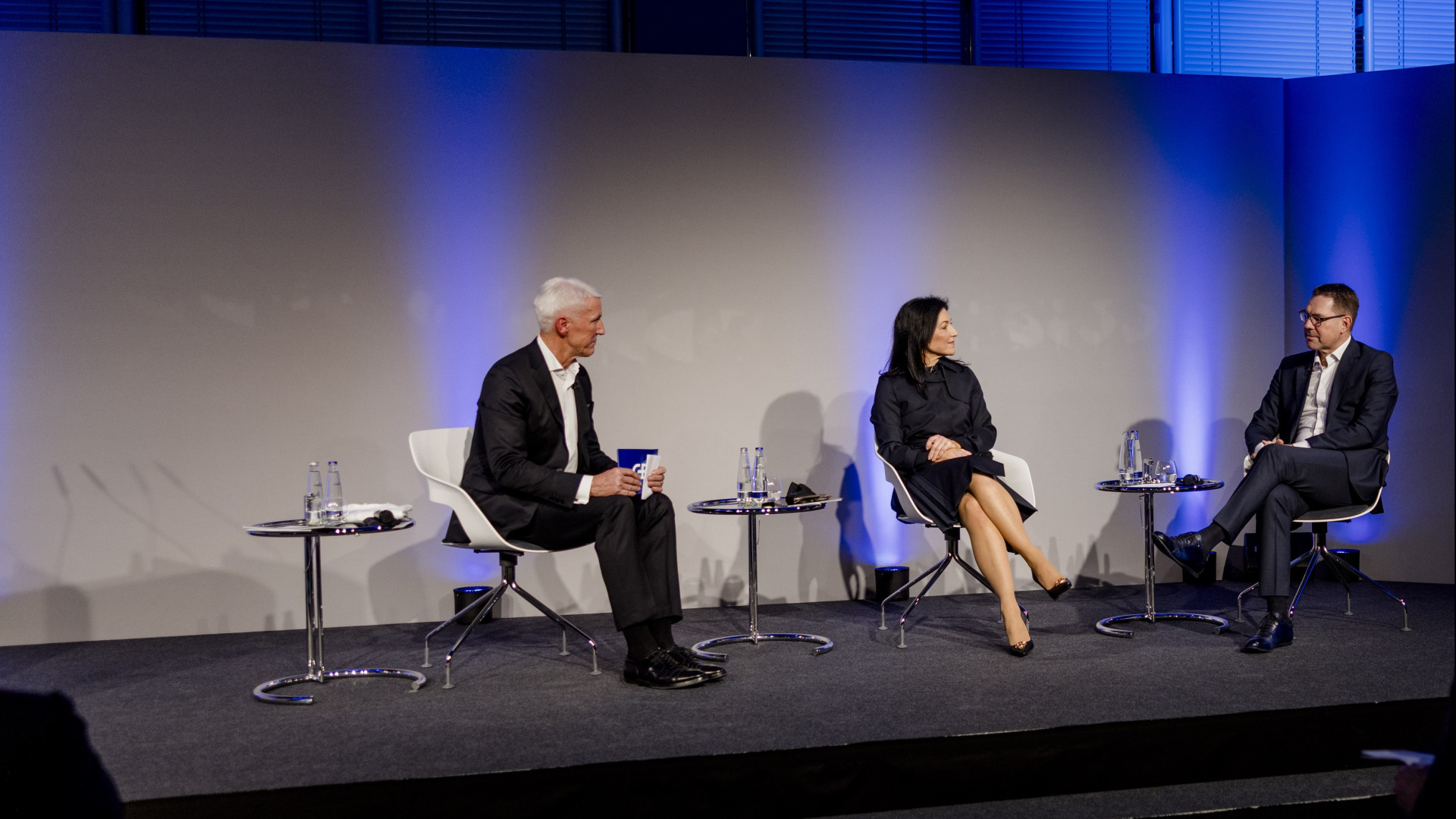 Drei Personen auf einer Bühne: Mann im Anzug mit Mikrofon, Frau im Kleid, Mann im Anzug, alle mit Wassergläsern.