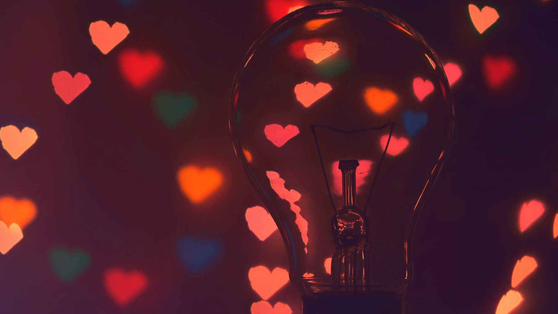 Transparente Glühbirne im Vordergrund, unscharfe, leuchtende Herzen in Rot, Pink, Blau und Gelb im Hintergrund.