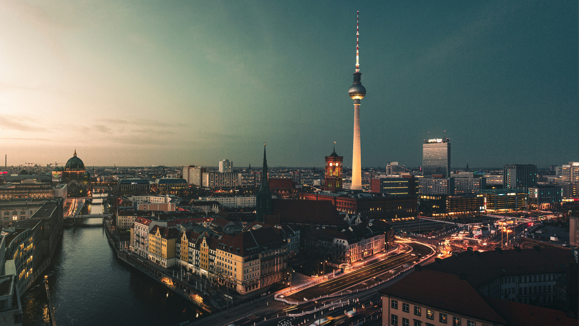 Stadtansicht von Berlin bei Dämmerung mit Fluss, Kuppel, Fernsehturm und beleuchteten Straßen. Keine Personen sichtbar.