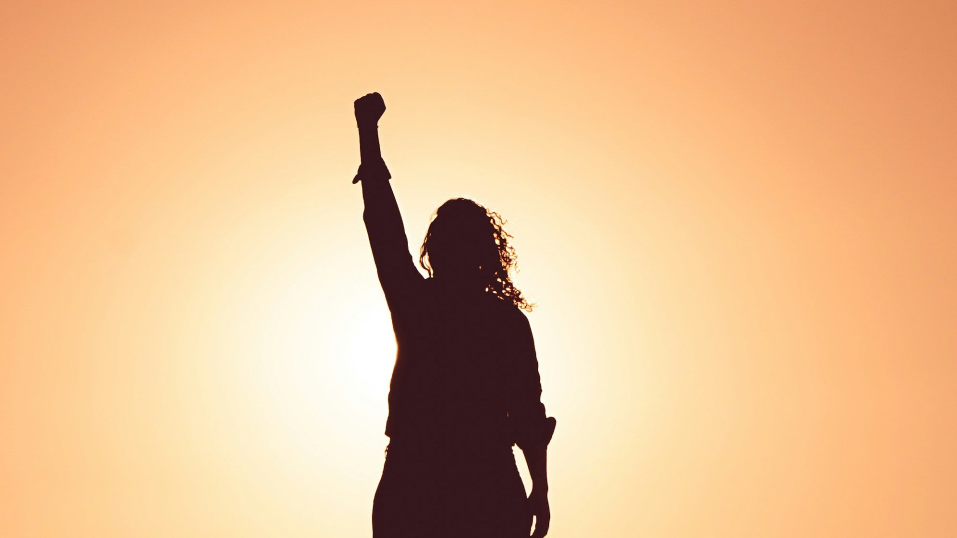 Silhouette einer Frau mit erhobener Faust vor einem orangefarbenen Sonnenuntergangshintergrund.