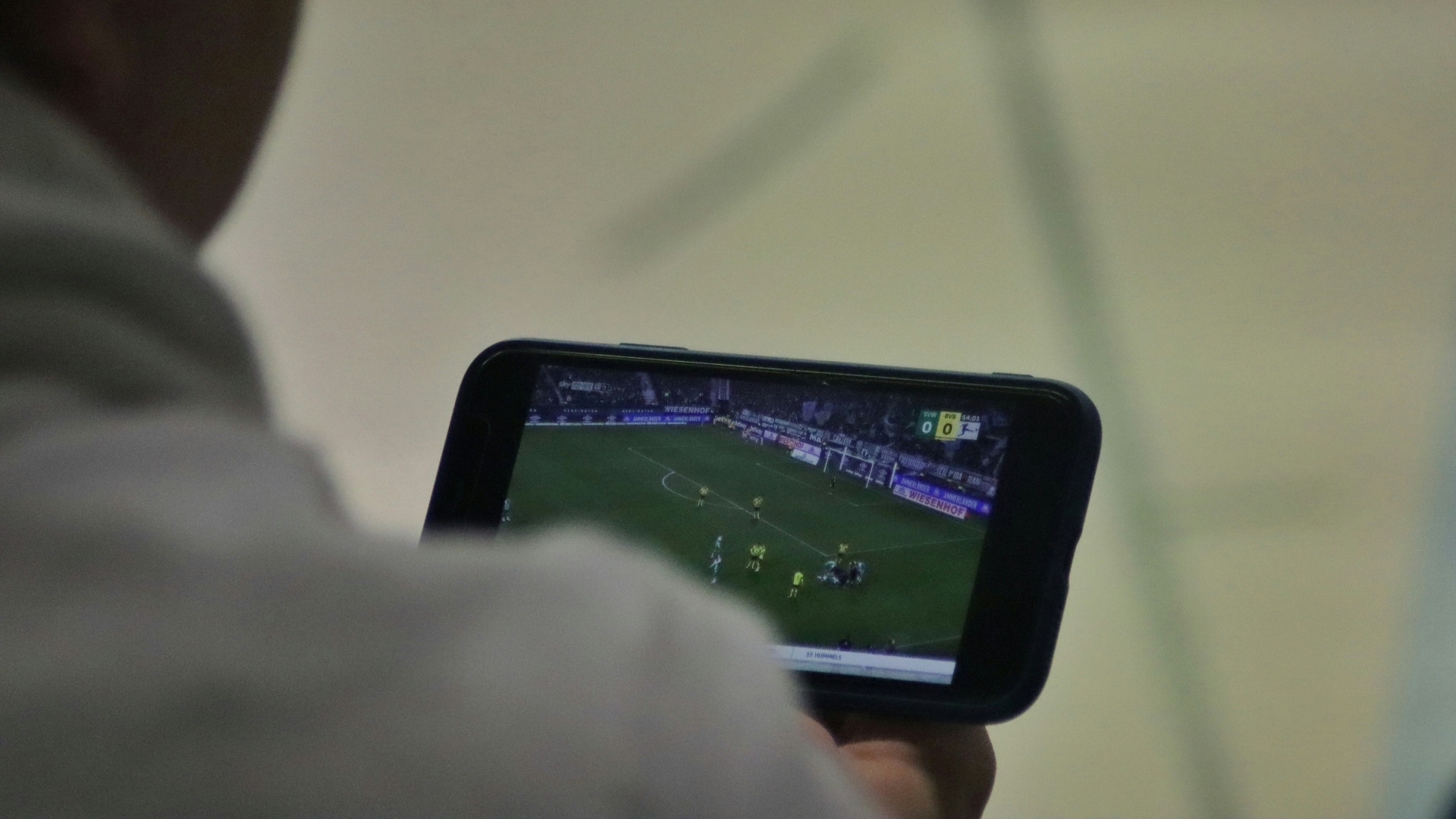 Mann mit hellem Pullover schaut auf Smartphone-Bildschirm mit Fußballszene und Punktestand 0:0.