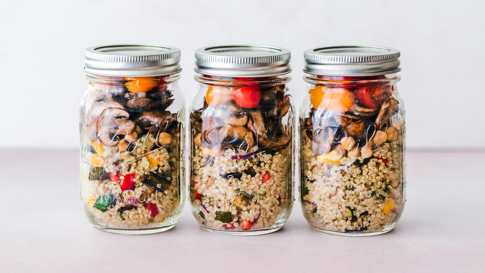 Drei Schraubgläser mit Quinoa, buntem Gemüse und kleinen Tomaten auf einer flachen Oberfläche. Metallene Schraubverschlüsse.