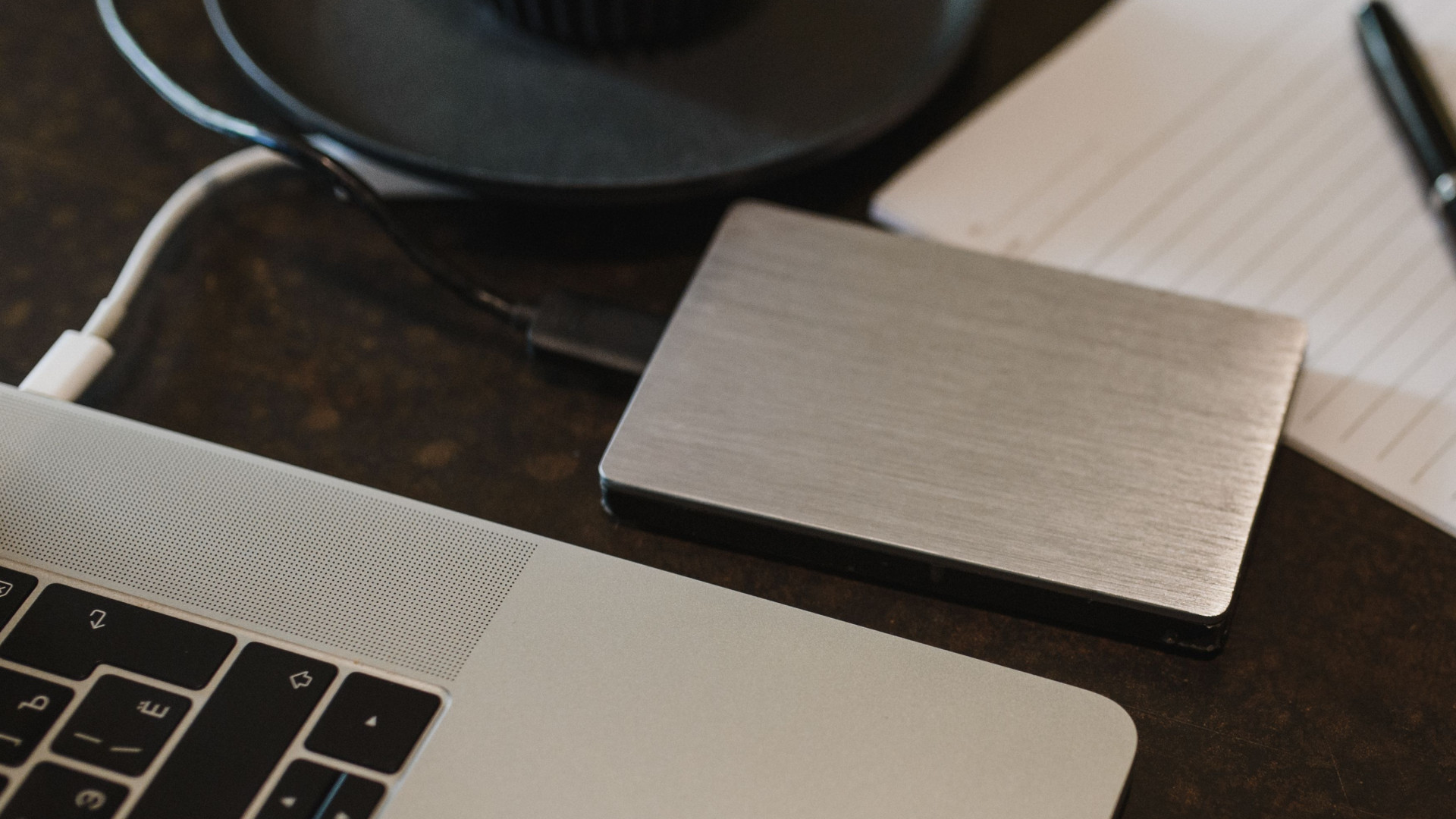 Externe Festplatte, Laptop, USB-Kabel, Kaffeebecher, Blatt Papier und Stift auf einem dunklen Tisch.