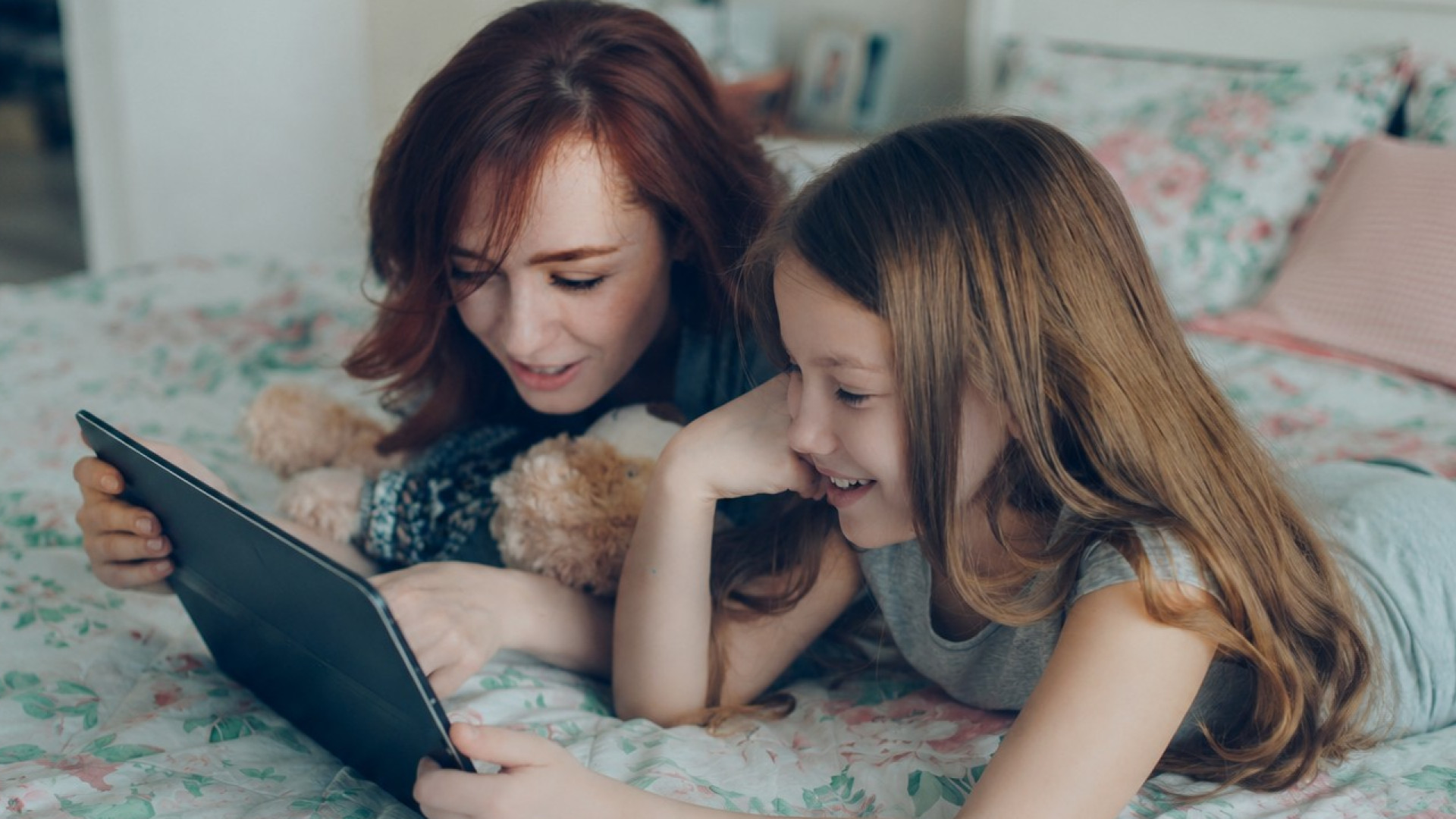 Mutter und Kind liegen auf einem Bett, schauen auf ein Tablet. Kind hat einen Teddybären neben sich.