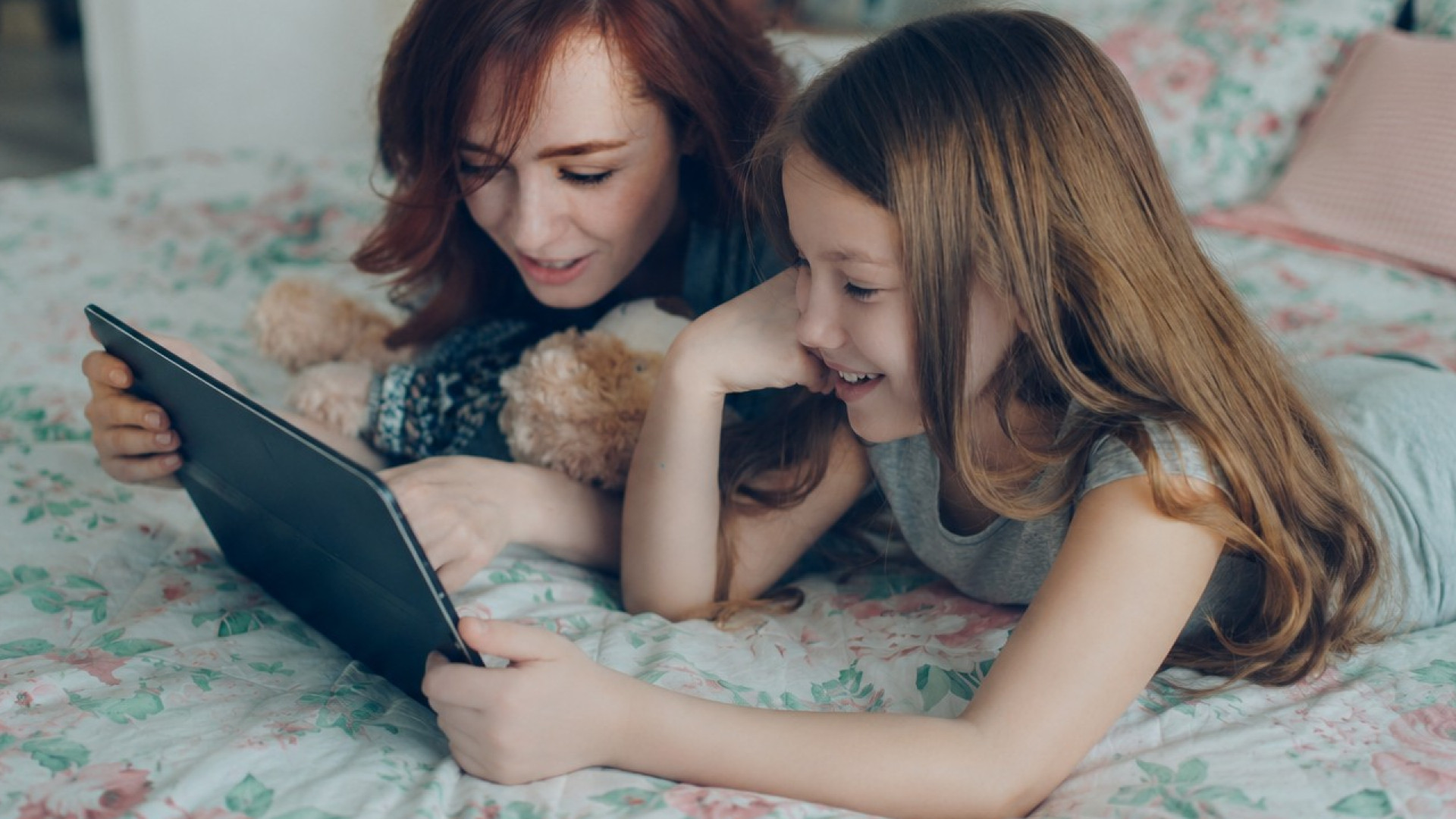 Mutter und Kind liegen auf einem Bett, schauen auf ein Tablet. Kind hat einen Teddybären neben sich.