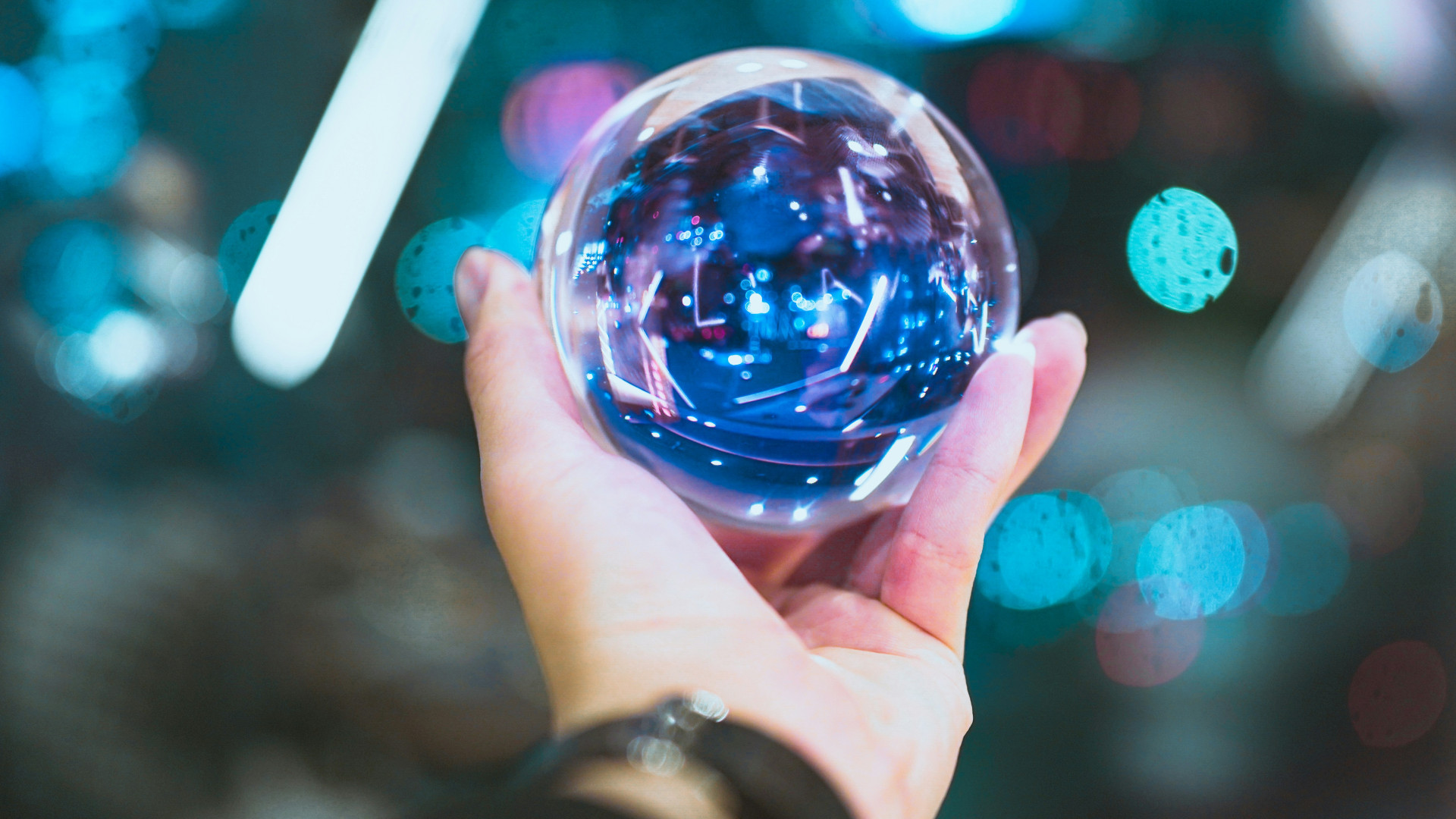 Eine Hand hält eine transparente Glaskugel mit reflektierenden blauen und lilafarbenen Lichtern. Unscharfe Lichtpunkte im Hintergrund.
