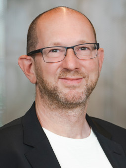 Andreas Streim, Person mit Brille und Bart, trägt schwarzen Blazer über weißem Oberteil vor unscharfem Hintergrund.