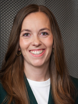 Frau mit langen, braunen Haaren lächelt in dunkelgrünem Blazer vor metallischer, strukturierter Oberfläche.