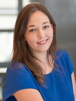 Person mit langen, braunen Haaren, blauem Oberteil und schwarzen Hosen, lächelt in die Kamera. Unscharfe Fenster im Hintergrund.