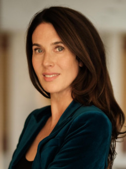 Frau in dunkelblauem Anzug mit verschränkten Armen, lächelt und blickt in die Kamera, unscharfer heller Innenraum im Hintergrund.