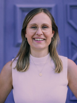 Person mit langen, glatten Haaren in ärmellosem Oberteil vor lila Wand mit geometrischen Mustern, lächelt in die Kamera.