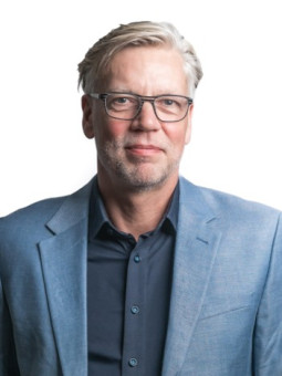 Mann mit grauen Haaren und Brille in hellblauem Blazer vor weißem Hintergrund, neutraler Gesichtsausdruck.