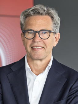 Marcel de Groot, grauhaariger Mann in Anzug, vor rotem Vodafone-Logo, Arme verschränkt, leicht lächelnd.