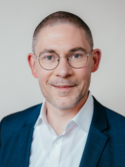 Mann mit kurzen grauen Haaren, Brille, dunkelblauem Anzug und weißem Hemd, lächelt in die Kamera.